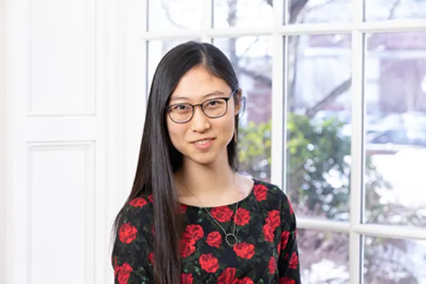 A young woman with glasses stands in front of a window, light illuminating her face and creating a warm atmosphere.