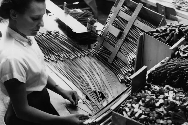 Factory worker organizes metal rods and components on an assembly table surrounded by bins.