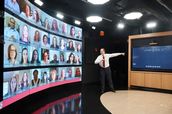 Instructor gestures in a virtual classroom with a large video wall of participants and a digital chalkboard