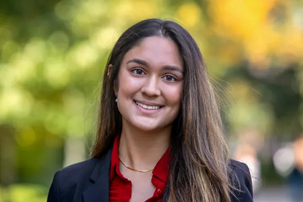 A young woman wearing a red shirt and a black blazer, standing confidently with a neutral expression.