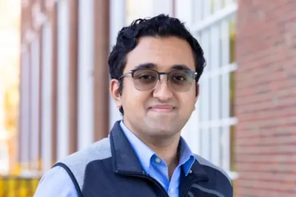 Individual wearing glasses, a blue shirt, and a gray vest, standing in front of a brick building with large windows.