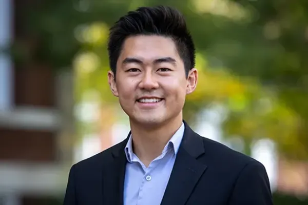 Person with short dark hair wearing a black blazer and light blue shirt, smiling outdoors with trees in background.