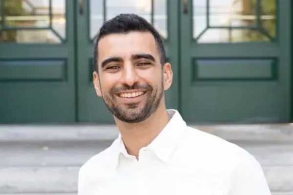 Person with short dark hair and beard wearing a white shirt, smiling in front of green double doors.