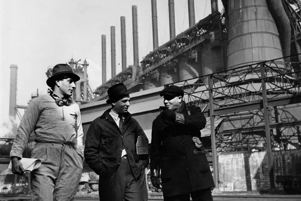 Three industrial workers talk while walking outside a steel mill with tall smokestacks.