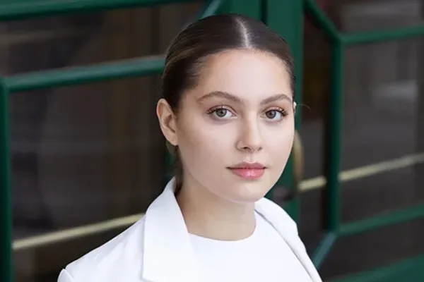 Person in white blazer looking at the camera in front of green-framed glass doors.