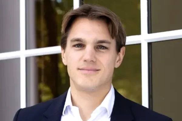 Person with short brown hair in a dark blazer and white shirt standing in front of a window.