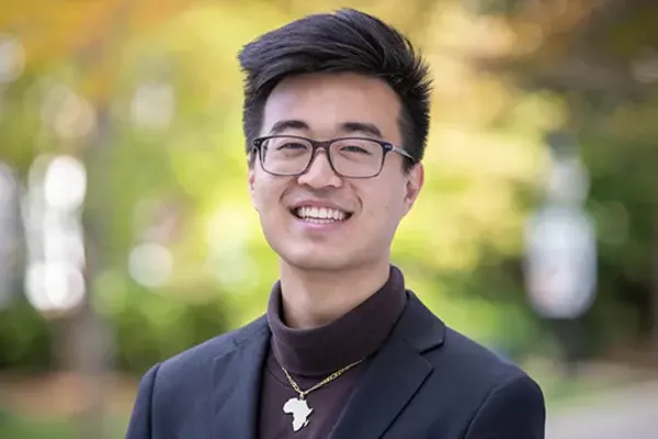 Person with short, styled dark hair and glasses smiling outdoors, wearing a dark blazer and Africa-shaped necklace.