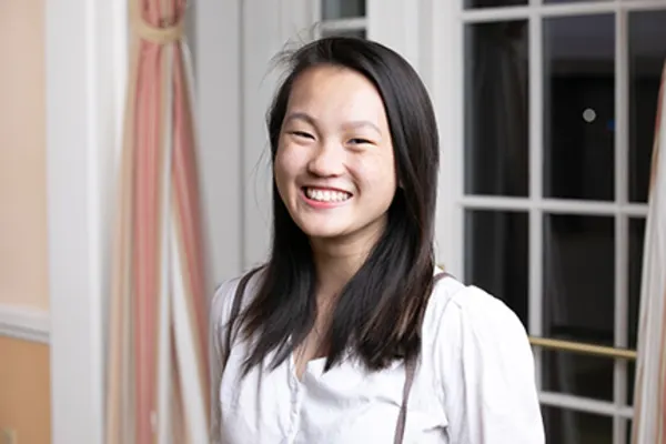 A young woman smiling brightly in front of a window, with natural light illuminating her face.