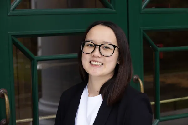 Person with straight dark hair and glasses, wearing a black blazer and white shirt, smiling while standing in front of green framed glass doors.