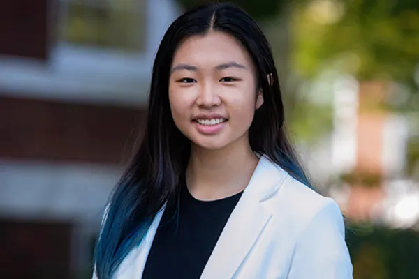 A young woman with blue hair wearing a white blazer, standing confidently with a neutral expression.