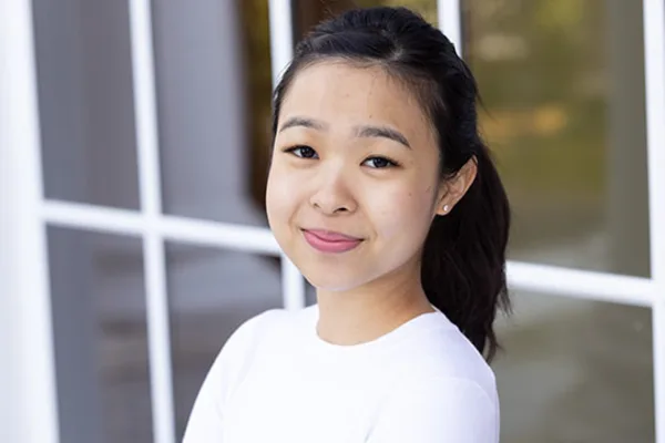 A young Asian woman stands in front of a window, with natural light illuminating her features.