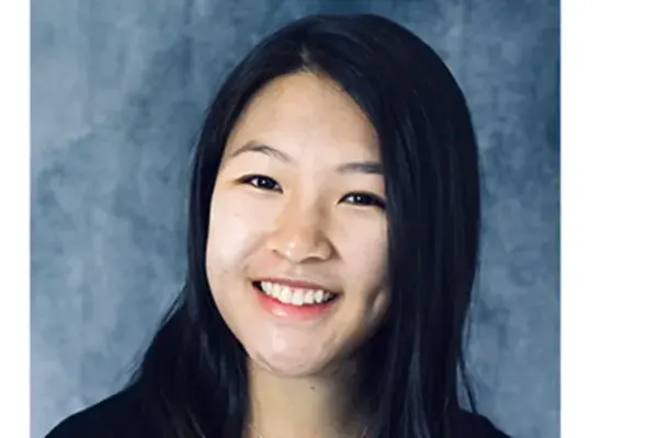 Smiling individual with long dark hair, wearing a black top, posed in front of a blue-gray textured backdrop.