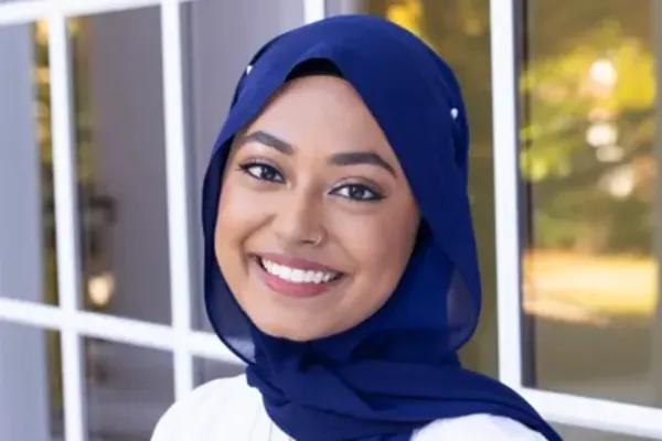 Smiling individual wearing a navy blue headscarf and white top, standing in front of a windowed building.