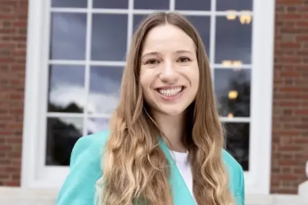 Person with long wavy hair wearing a teal blazer, smiling in front of a large window with brick framing.