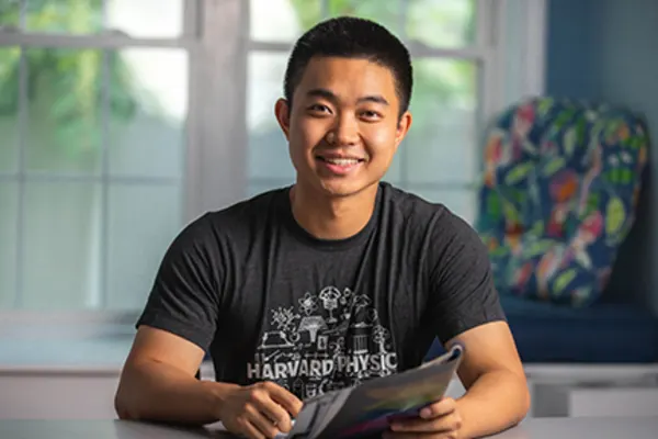 A young man sits at a table holding a magazine, smiling at the camera. He wears a dark "Harvard Physics" t-shirt. A window and a colorful chair are in the background.