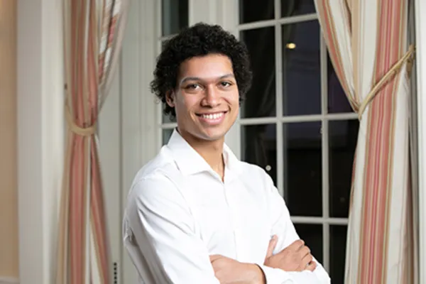 Man in a white shirt standing, looking directly at the camera with a neutral expression.