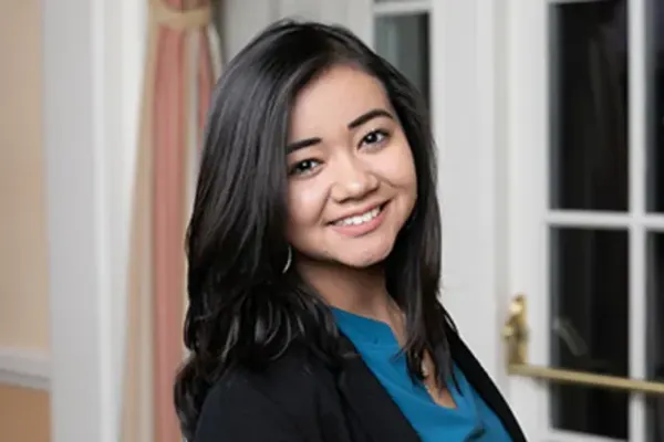 Person with shoulder-length dark hair wearing a teal top and black blazer, smiling indoors near glass doors.