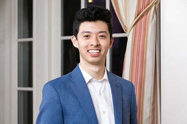 A young man wearing a blue suit and white shirt, standing confidently with a neutral expression.