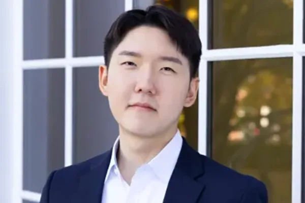 Individual with short dark hair in a navy suit and white shirt, standing in front of a window with a blurred outdoor background.