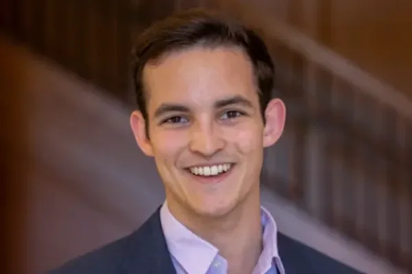 Smiling individual in a suit jacket and light purple shirt, standing in front of a blurred staircase background.