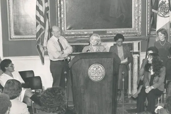 Muriel Siebert speaking at a podium with the NYC seal, surrounded by officials and attendees in a formal setting.