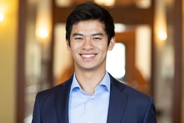 A young man in a blue shirt and jacket smiles warmly at the camera.