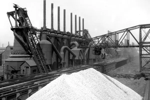 Large steel mill with smokestacks, conveyor belts, and a pile of raw limestone in the foreground.