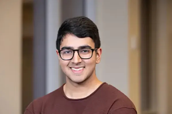 Person with short dark hair and glasses wearing a maroon shirt, smiling indoors with blurred background.