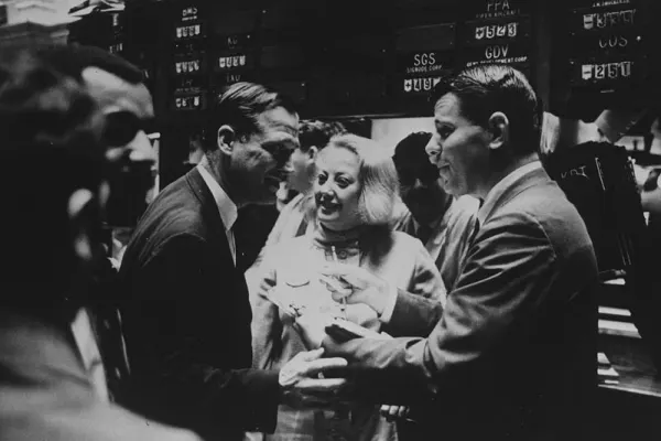 Muriel Siebert smiling among male colleagues on the NYSE trading floor, engaging in conversation.
