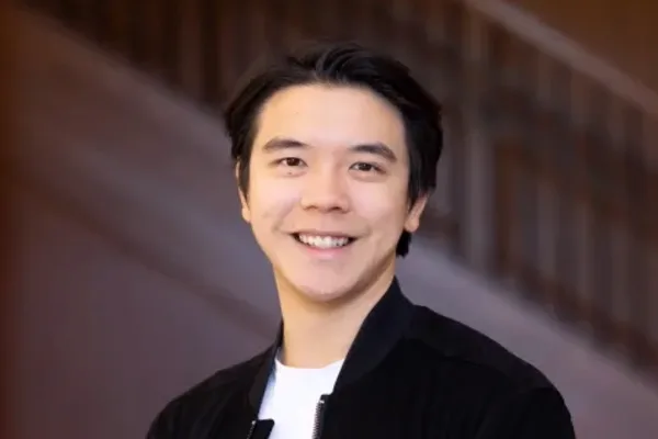 Person with short dark hair wearing a black jacket over a white shirt, smiling in front of a staircase background.