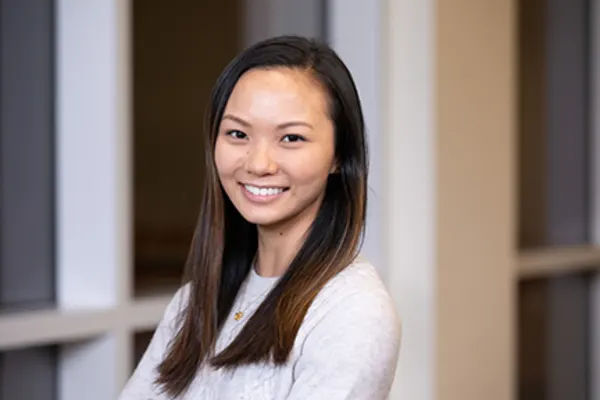 A woman with long hair wearing a white shirt, standing in a neutral background.