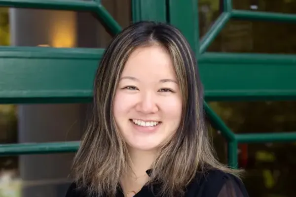 Smiling individual with shoulder-length hair and light highlights, wearing a black top, posed in front of a green-framed glass door.