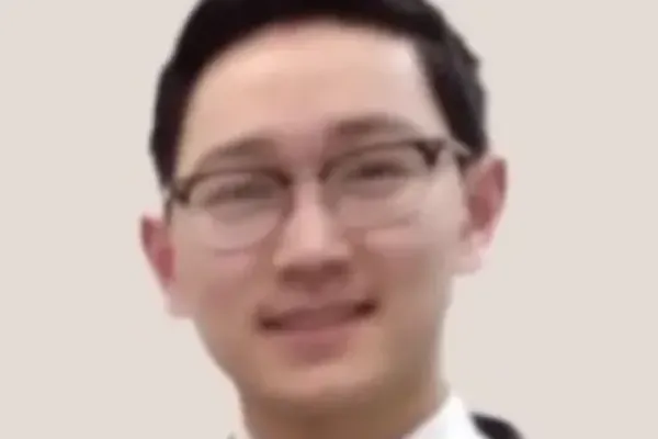 ChatGPT said: Individual wearing glasses and a suit with a white shirt and tie, standing against a plain light background.