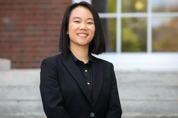 Smiling individual with straight dark hair in a black suit, standing outside in front of stone steps and a windowed brick building.