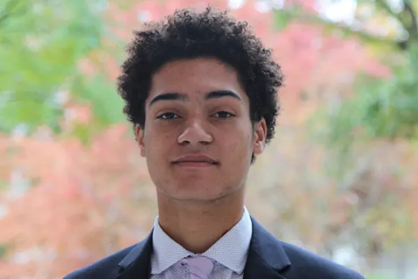 A young man in a suit and tie stands confidently in front of lush green trees.