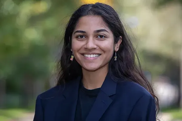 Person with long dark hair wearing a navy blazer and colorful earrings, smiling outdoors with greenery in the background.