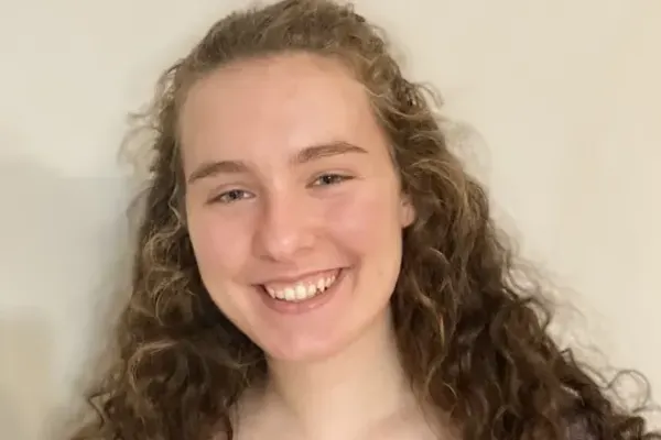 Person with long curly hair smiling in front of a plain light-colored wall.