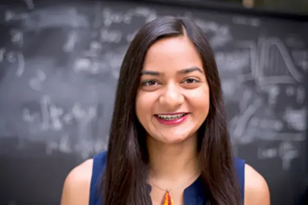 Person with long dark hair wearing a navy top, smiling in front of a chalkboard covered in math equations and graphs.