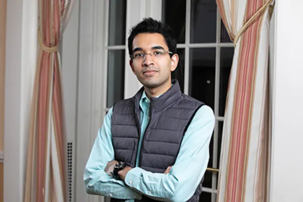 A man in a vest stands by a window, looking out at the view with a thoughtful expression.