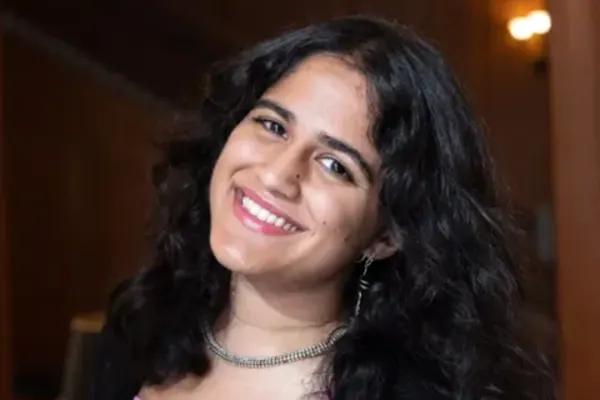 Smiling individual with wavy dark hair, wearing a necklace and pink top, standing in a warmly lit indoor setting.