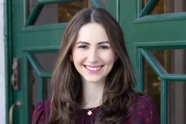 Smiling individual with long brown hair, wearing a dark red patterned top, standing in front of a green door with window panels.