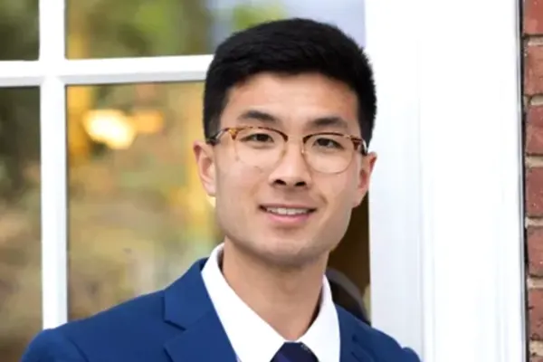 Person with short black hair and glasses wearing a blue suit, white shirt, and tie, standing in front of a window.