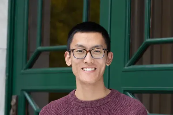 Person wearing glasses and a maroon sweater smiling in front of green-framed glass doors.