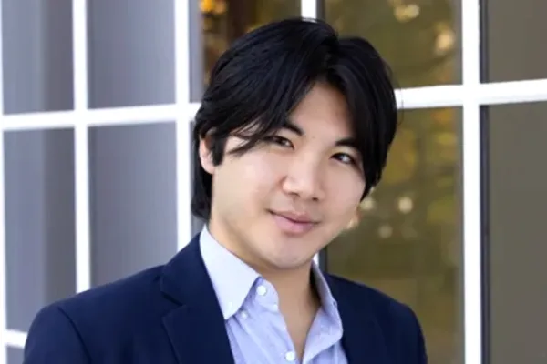 Individual with medium-length dark hair, wearing a blue blazer and light blue shirt, standing in front of a windowed building.