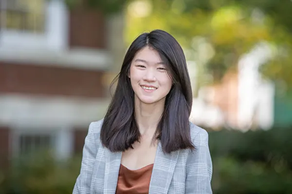 A young woman in a blazer and blouse stands outside, smiling confidently against a blurred urban background.