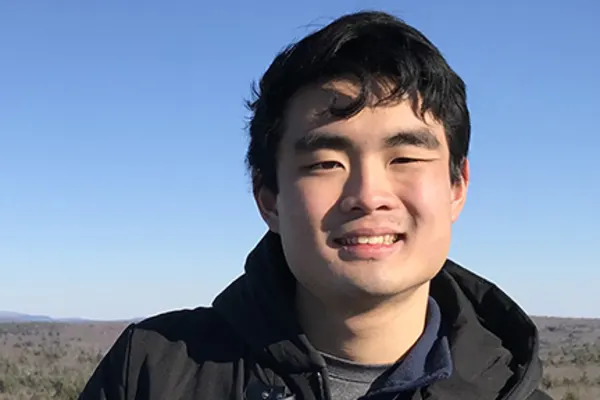 A person with short dark hair smiles outdoors, wearing a dark jacket. The background shows a clear blue sky and a landscape with trees and distant hills.