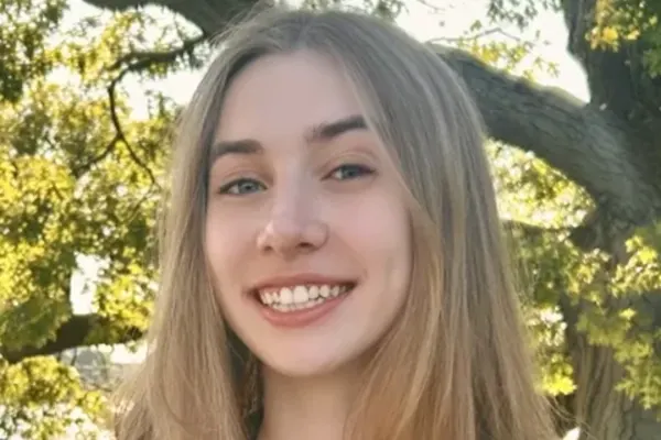 Person with long light brown hair smiling outdoors in front of leafy trees.
