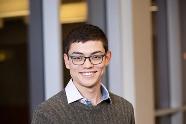 A young man with glasses and a sweater smiles warmly at the camera.