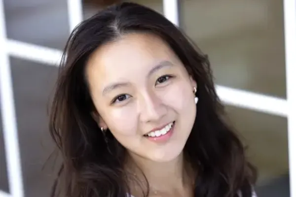 Smiling individual with long dark hair, wearing pearl earrings and a white top, standing in front of a window with a grid pattern.