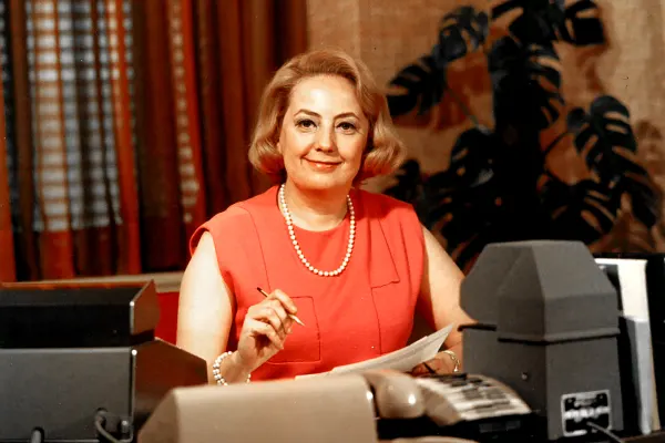 Muriel Siebert in a sleeveless coral dress sits at a desk with vintage office equipment, smiling and holding a pencil.